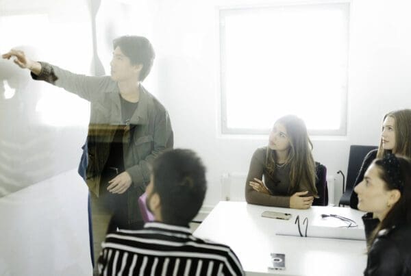 A group of students working together. One is presenting on the board, others are sitting around a table and watching.
