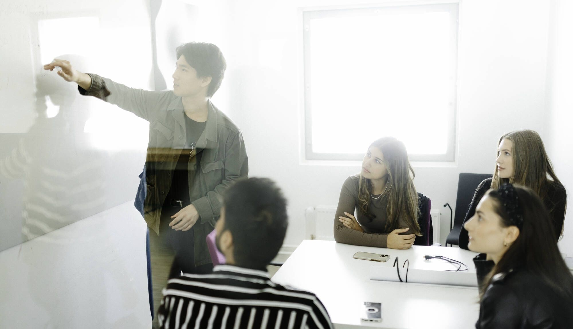 A group of students working together. One is presenting on the board, others are sitting around a table and watching.